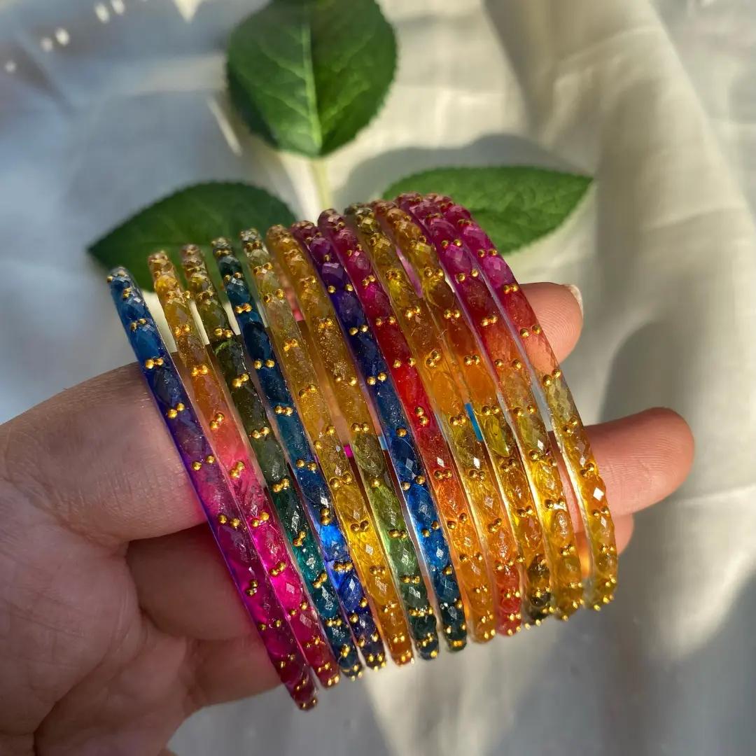 Fan of slim faceted glass bangles in deep pink, blue, amber and green with tiny gold bead dots held in a hand