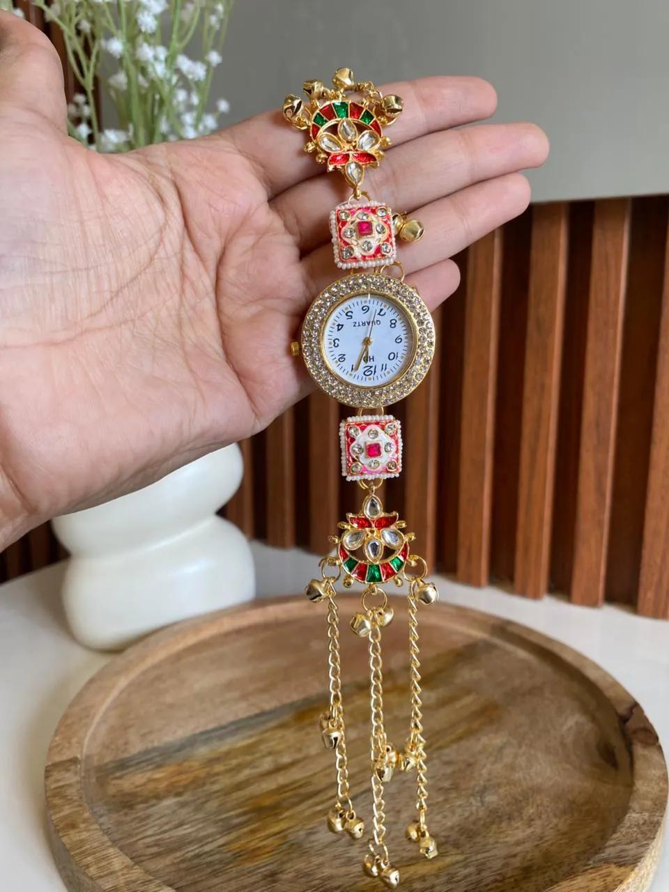Ornate gold-tone bracelet watch with kundan-style crown, red and green meenakari panels and ghungroo bell tassels