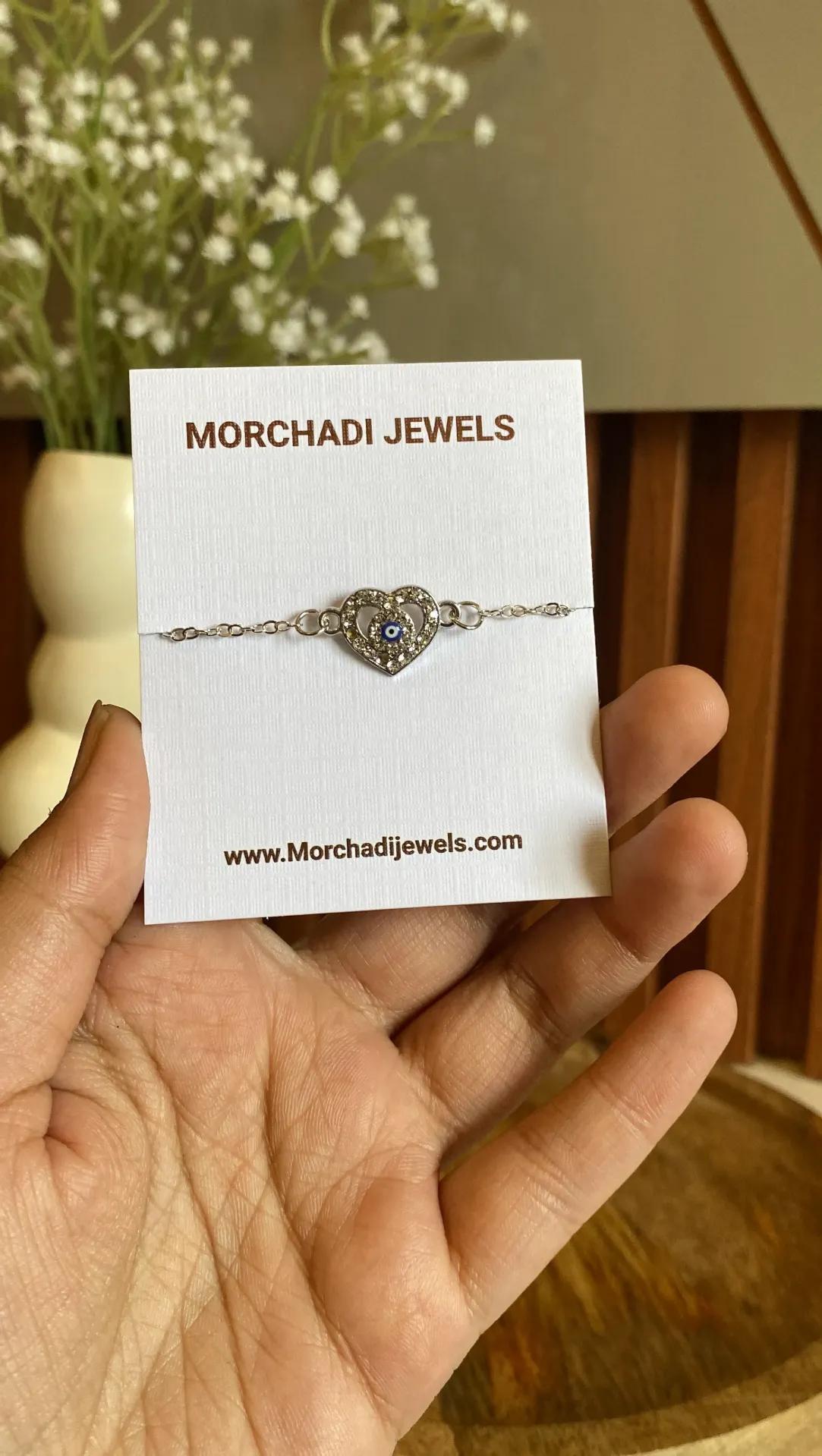 Silver-toned chain bracelet with a small stone-paved heart holding a blue evil eye bead, shown on a white card