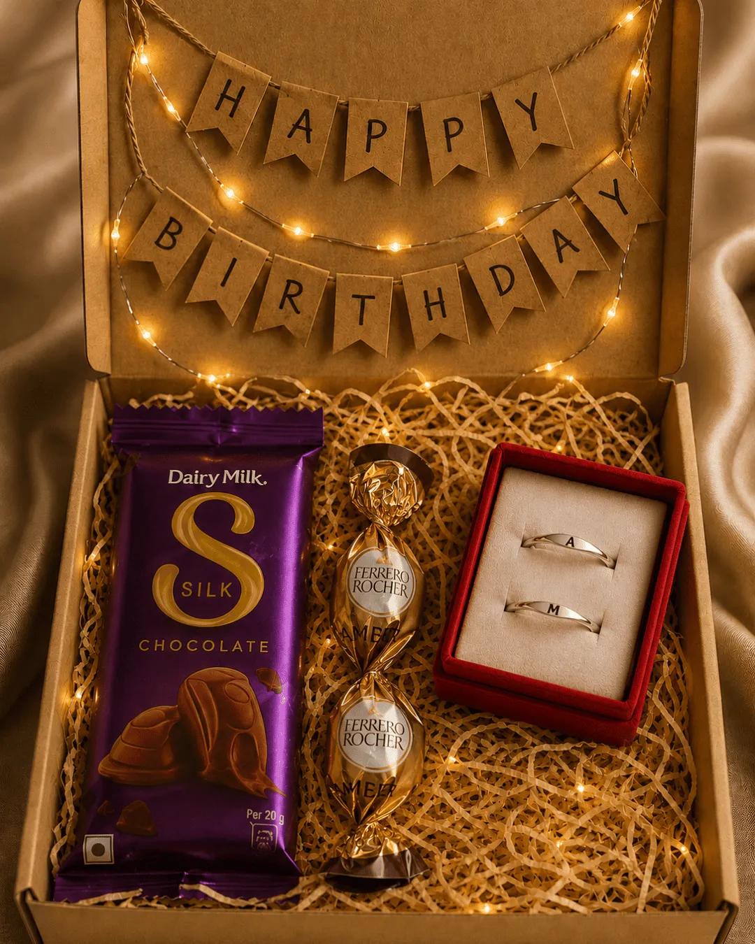 Silver-toned initial rings in a red velvet box beside chocolates and a birthday banner