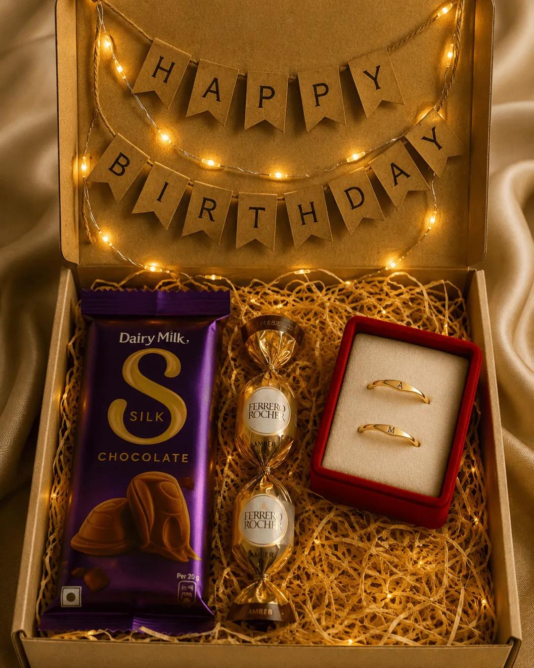 Gold-toned initial rings in a red velvet box beside chocolates and a birthday banner
