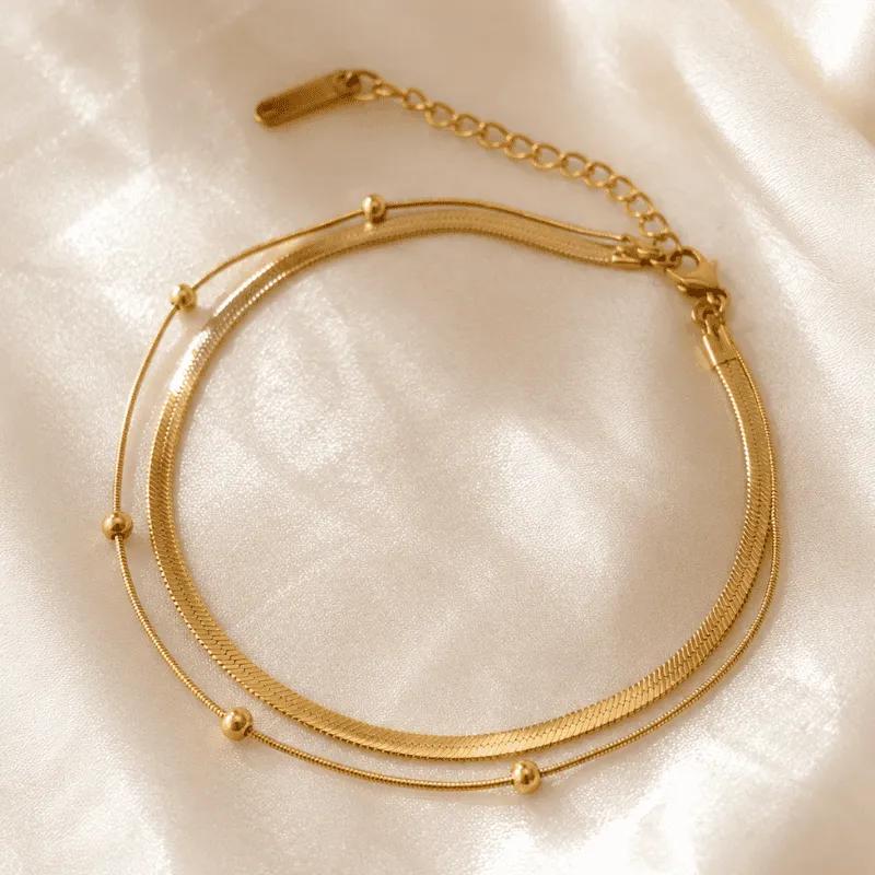 Gold-plated anklet with a flat herringbone chain and a fine bead-station chain sharing one extender clasp, on satin
